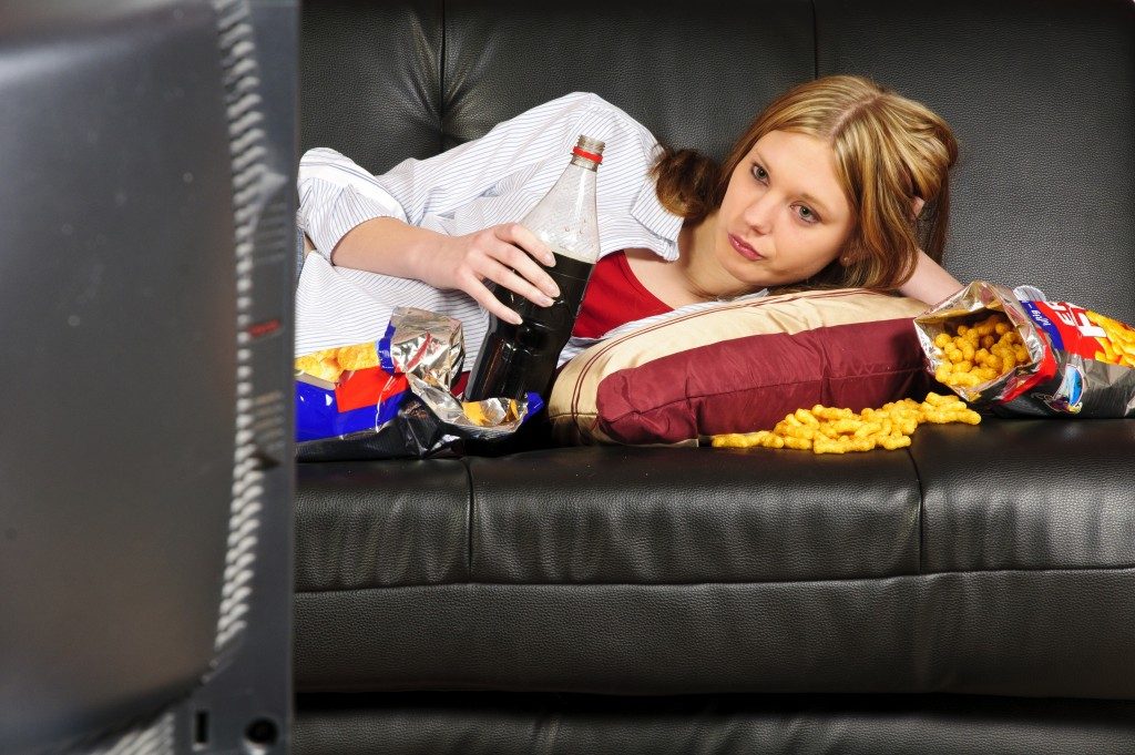 woman laying on the couch while eatinf junk foods and drinking softdrinks