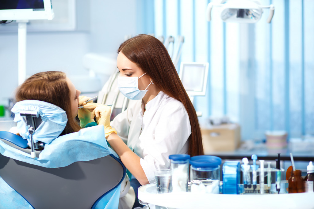 A dentist with a mask and gloves cleans out a patient's teeth while she is on a dentist's chair 