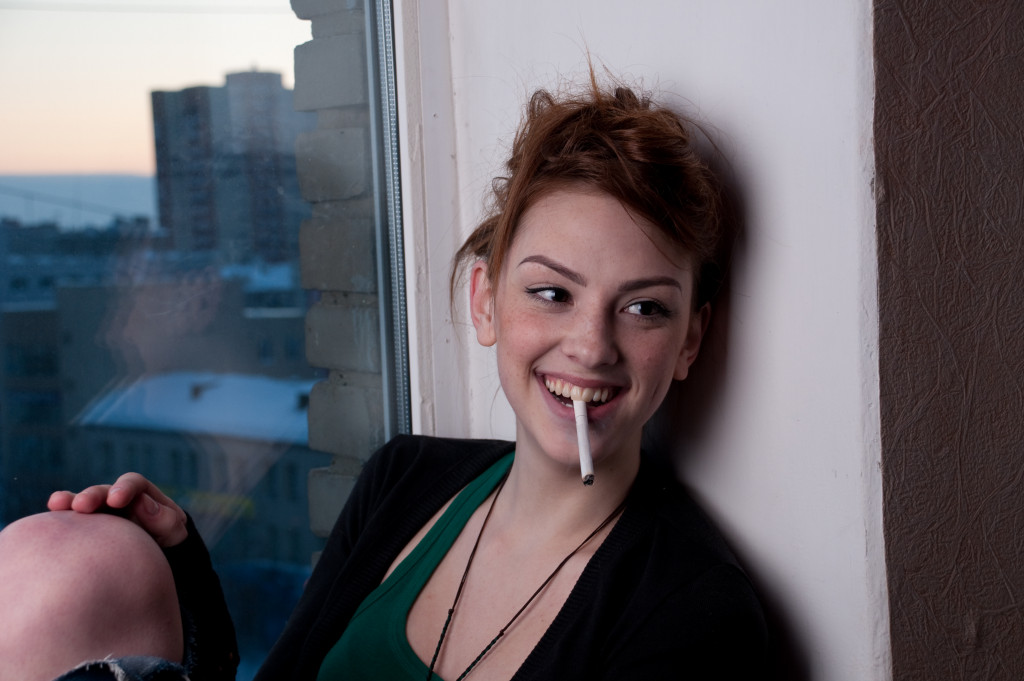 young woman in casual wear smoking a cigarette by the window