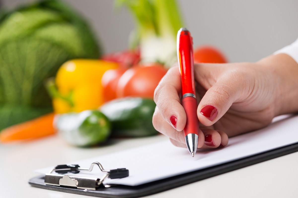 woman with red nail polish and red pen writing on a clipboard with vegetables on the side